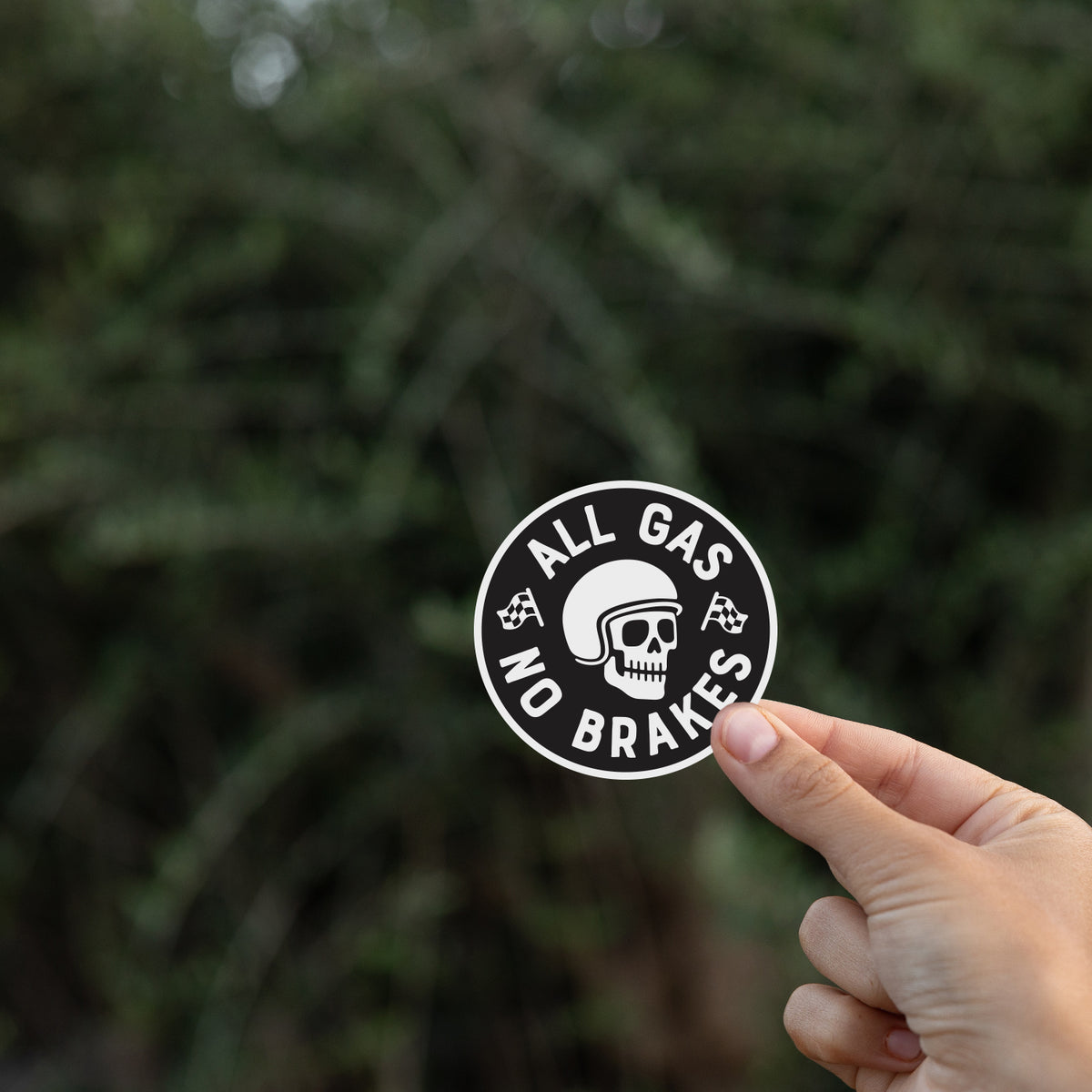 Fingers lift the All Gas No Brakes sticker against a dark blurred hedge of thin branches.