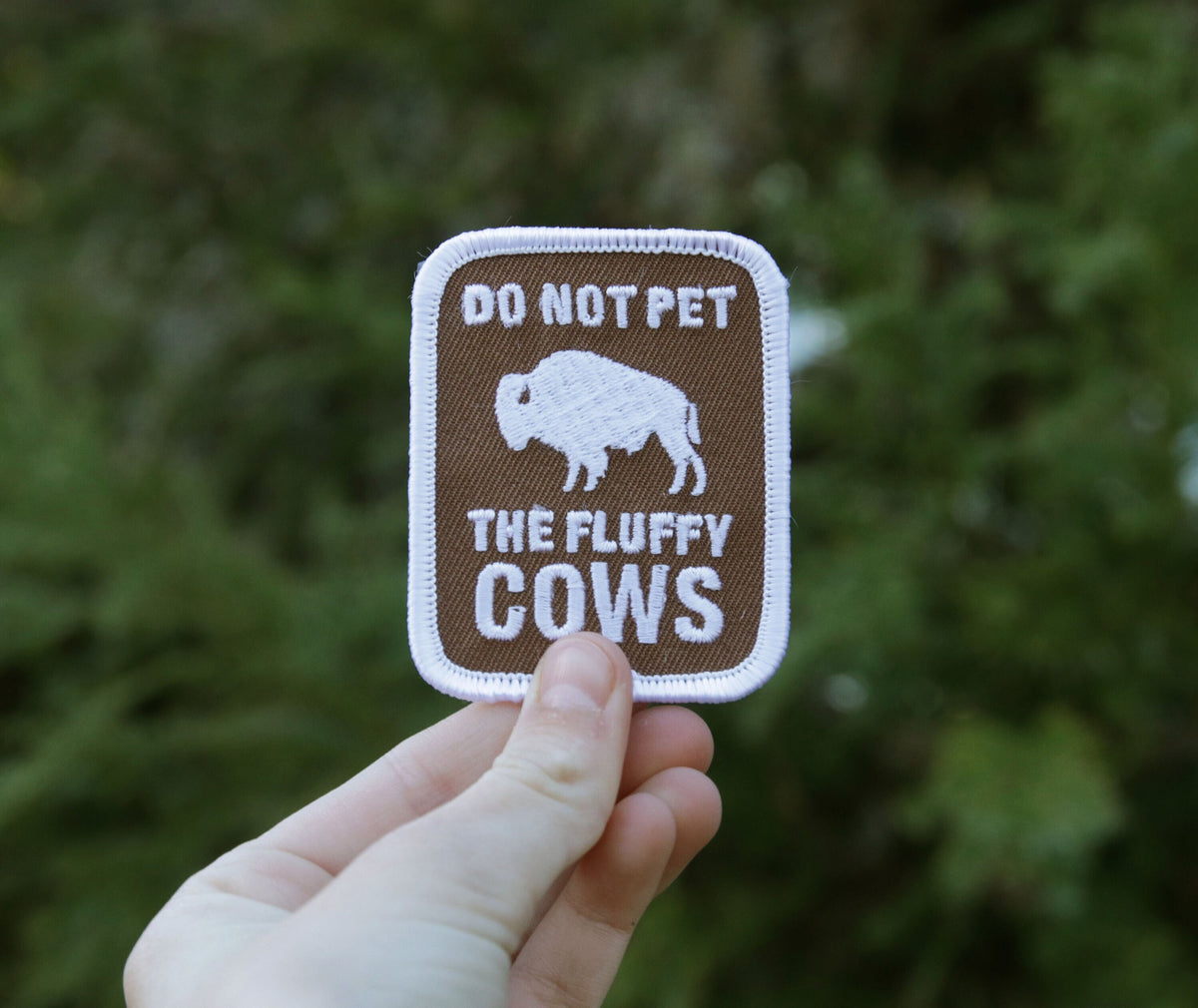 Fingertips hold the Don't Pet The Fluffy Cows patch further back, whole shape visible before dark shrubs.