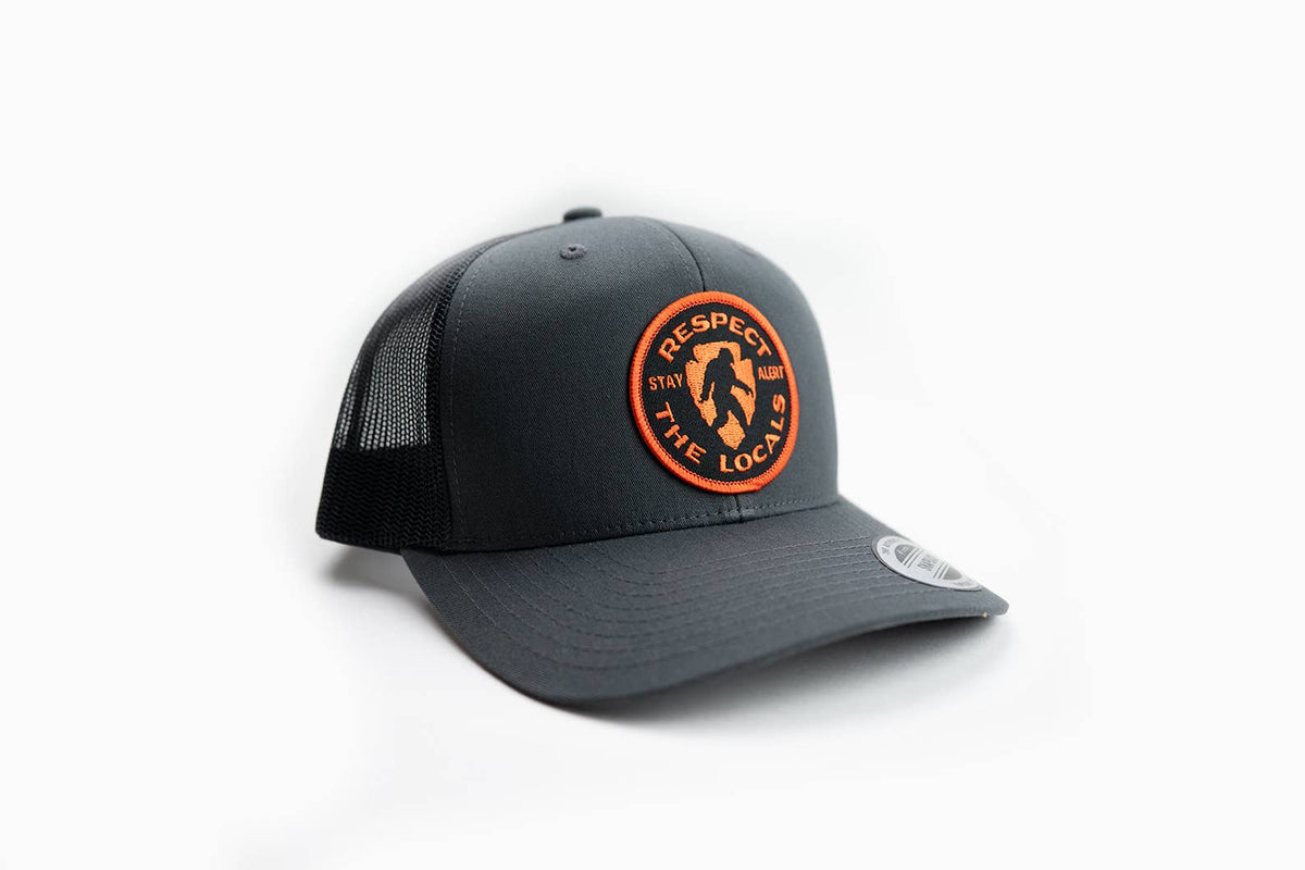 Stitched to the front of a charcoal trucker hat, the Respect the Locals Bigfoot patch sits above the brim.