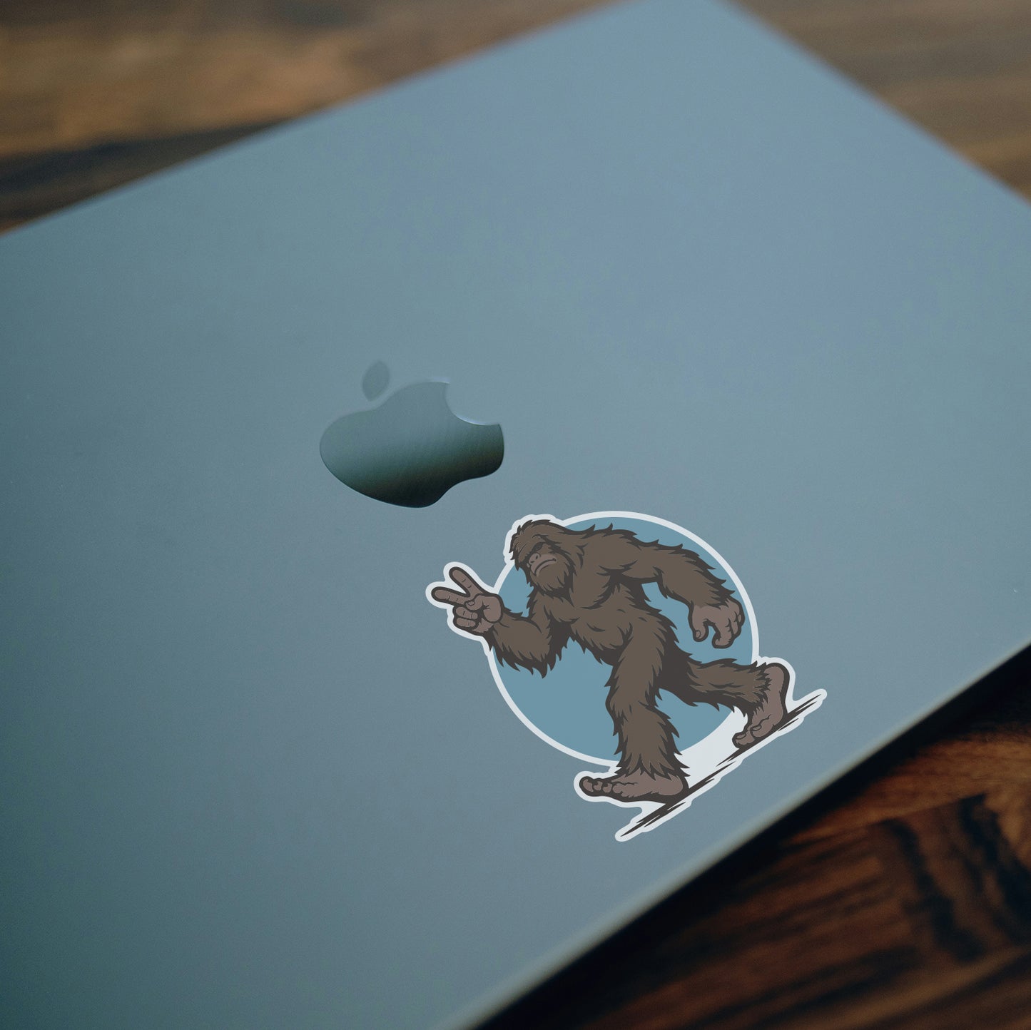 Stuck on a blue laptop lid above dark wood, the Bigfoot Peace Out sticker throws a two finger salute.