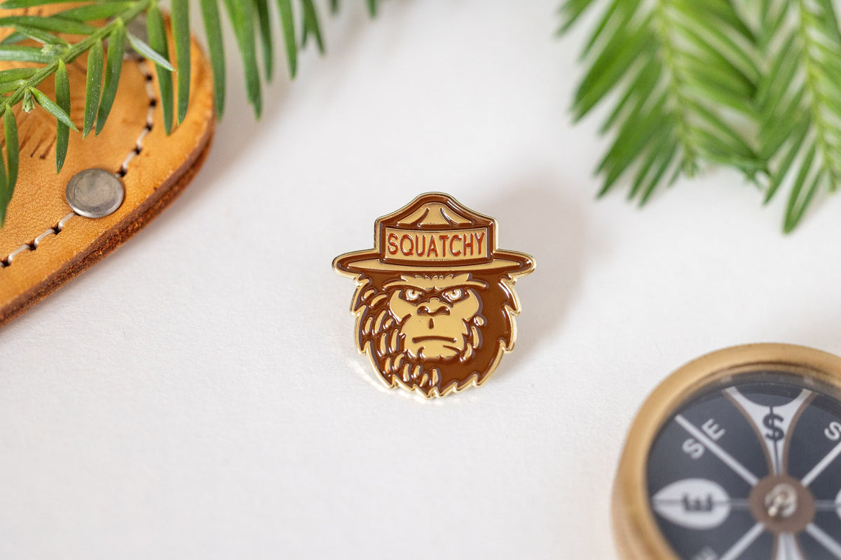 Off the card and on white, the Squatchy ranger Bigfoot enamel pin sits near pine and a leather flap.