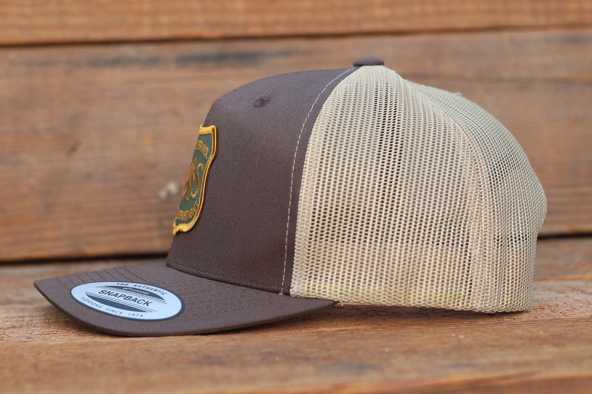From the side, the brown trucker hat shows deep tan mesh and the curved brim label on grey wood.