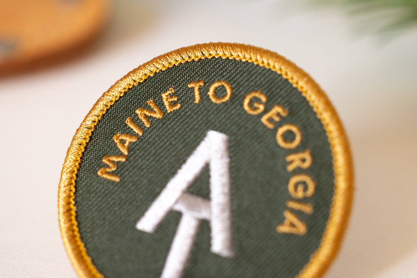 Close crop on the AT Maine to Georgia patch, where gold letters curve above the white trail monogram.