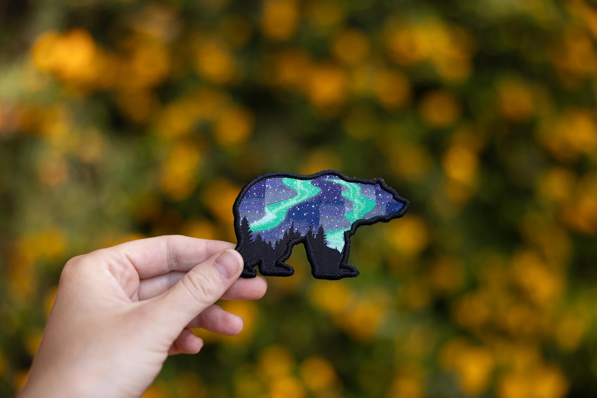 Closer to the golden blooms, fingers tilt the Northern Lights Grizzly Bear patch toward the camera.