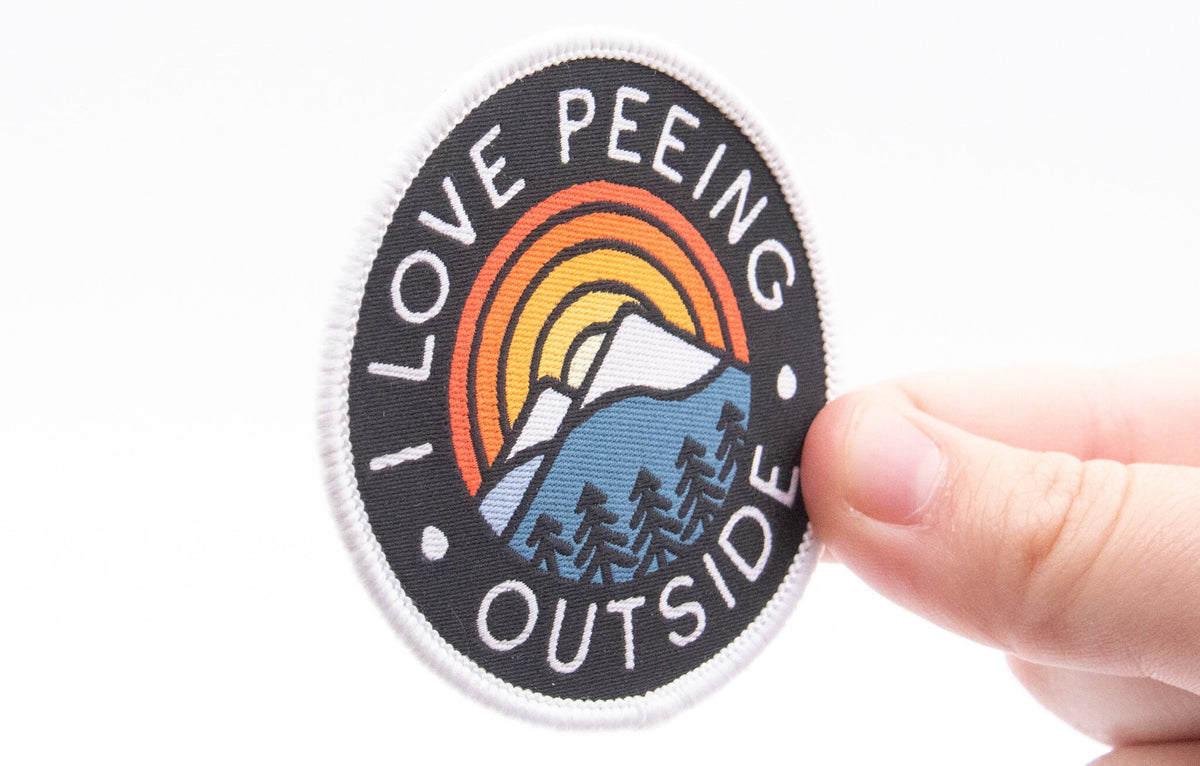 Fingers grip the edge of the I Love Peeing Outside patch, close enough to read every stitch in the border.