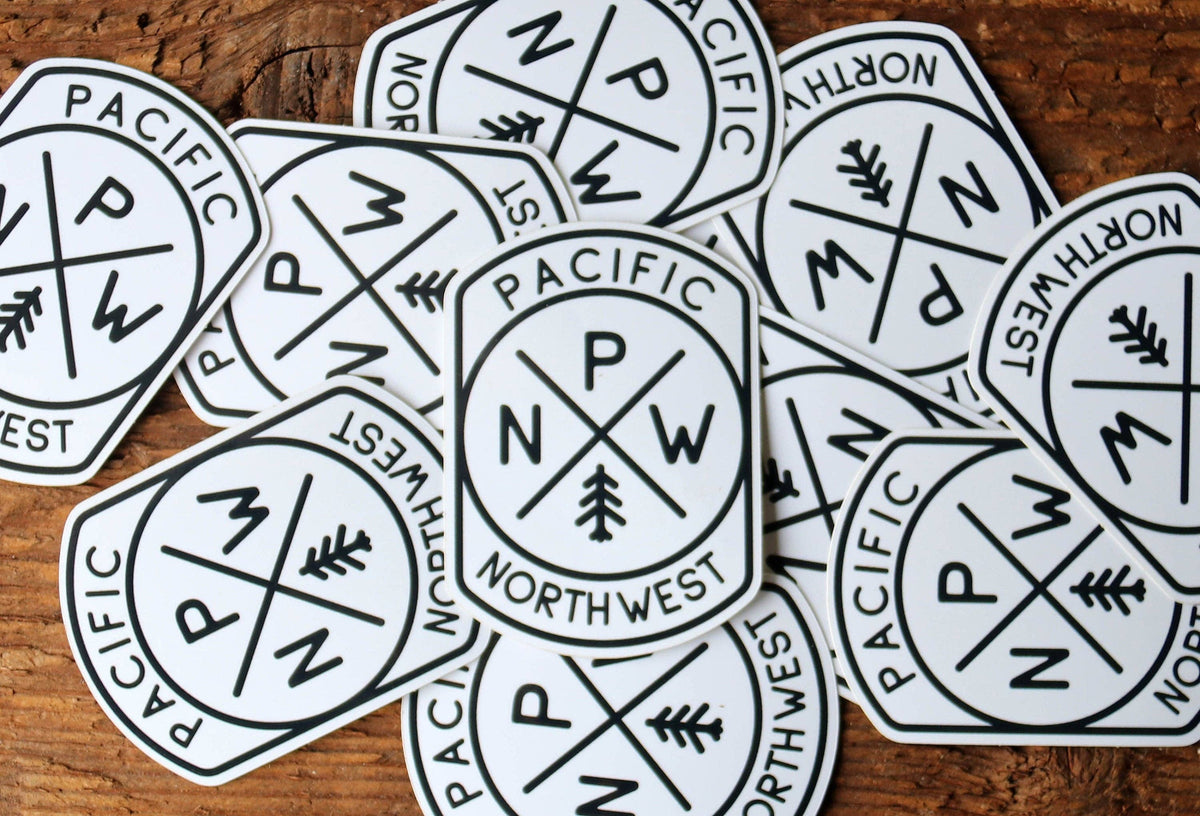 A scattered pile of Pacific Northwest Compass stickers lies face up on weathered wood, one badge centered upright.