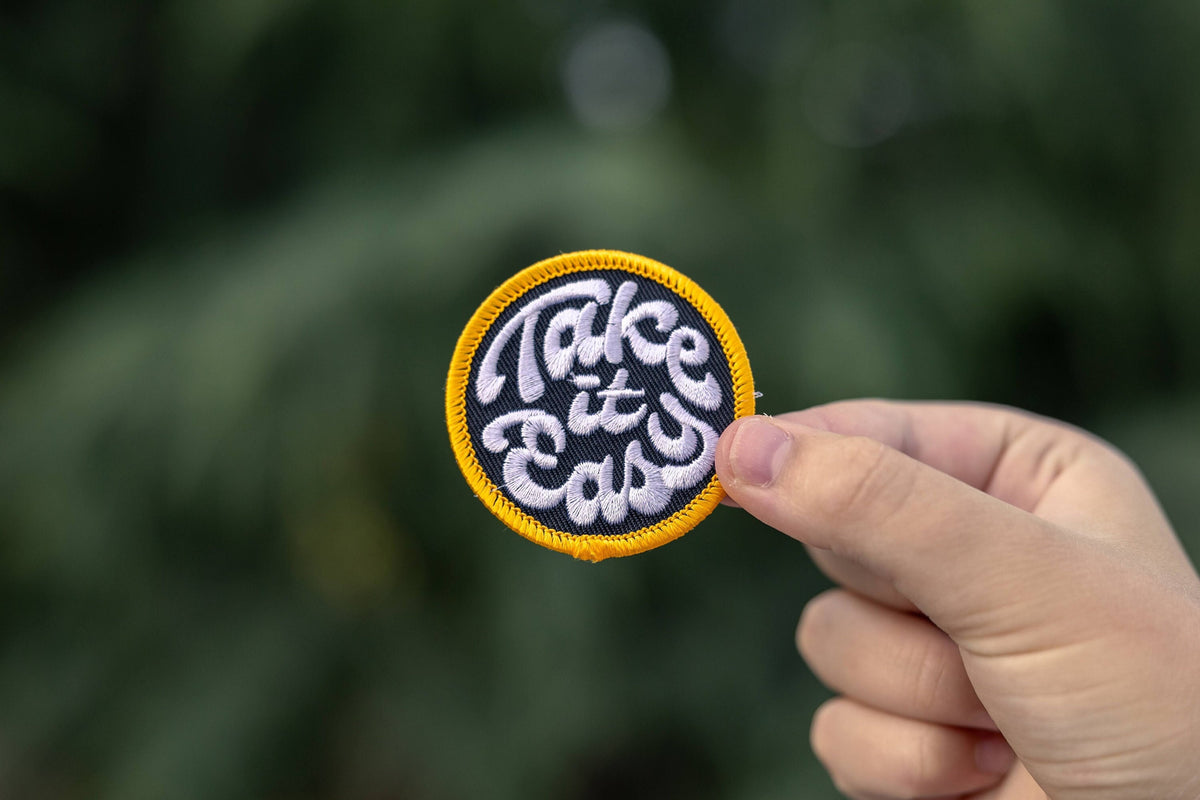 Fingers hold up the Take it Easy patch with green foliage blurred behind it.
