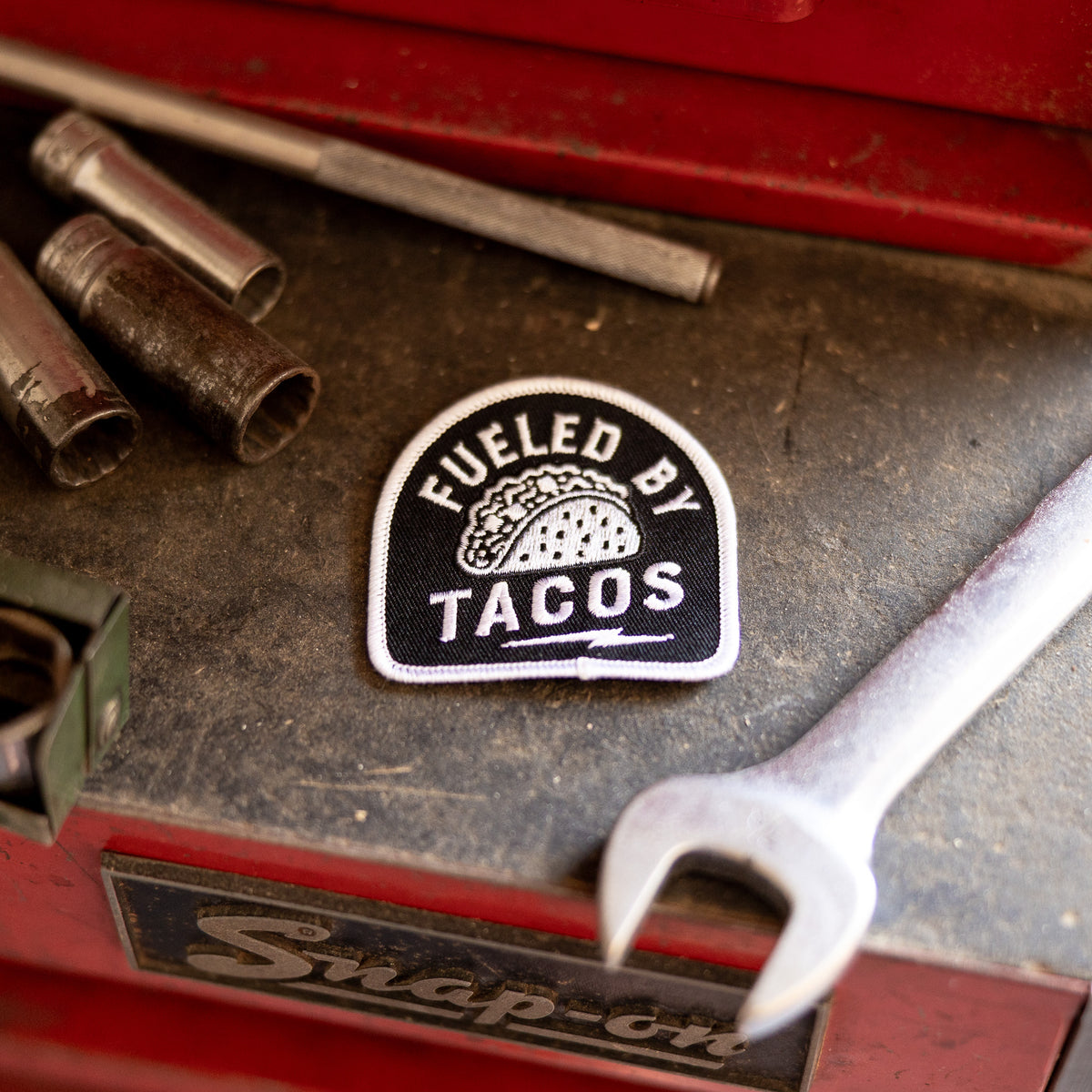 Set against worn tool steel, the Fueled By Tacos patch rests near a wrench and a row of socket bits.