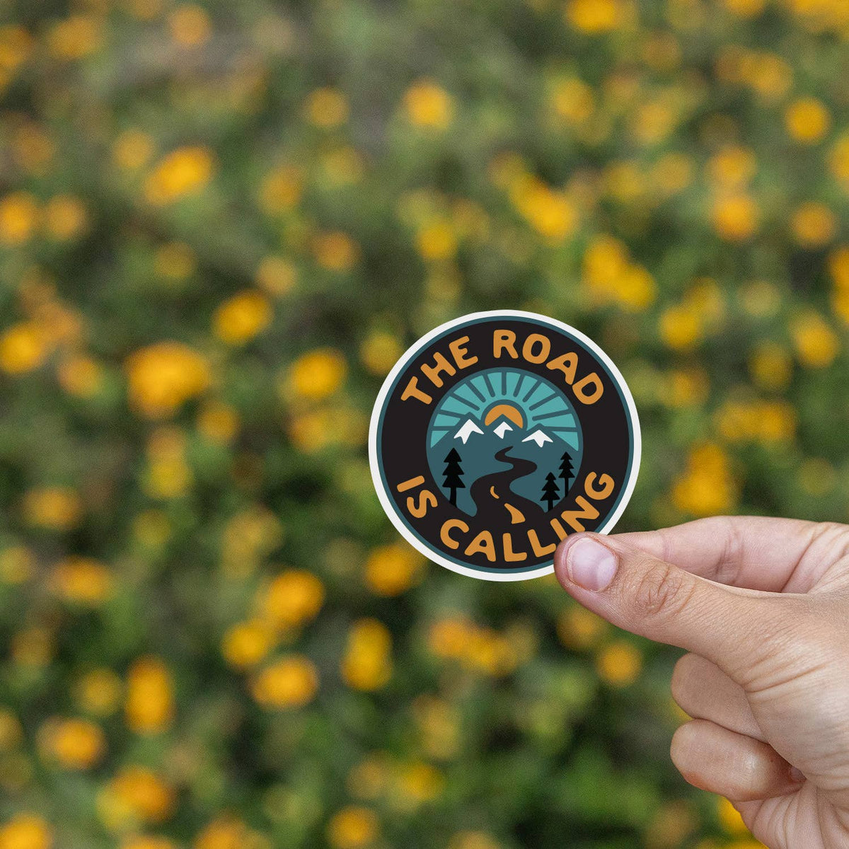 Held up over a field of yellow blossoms, the Road is Calling sticker keeps its teal and orange colors sharp.