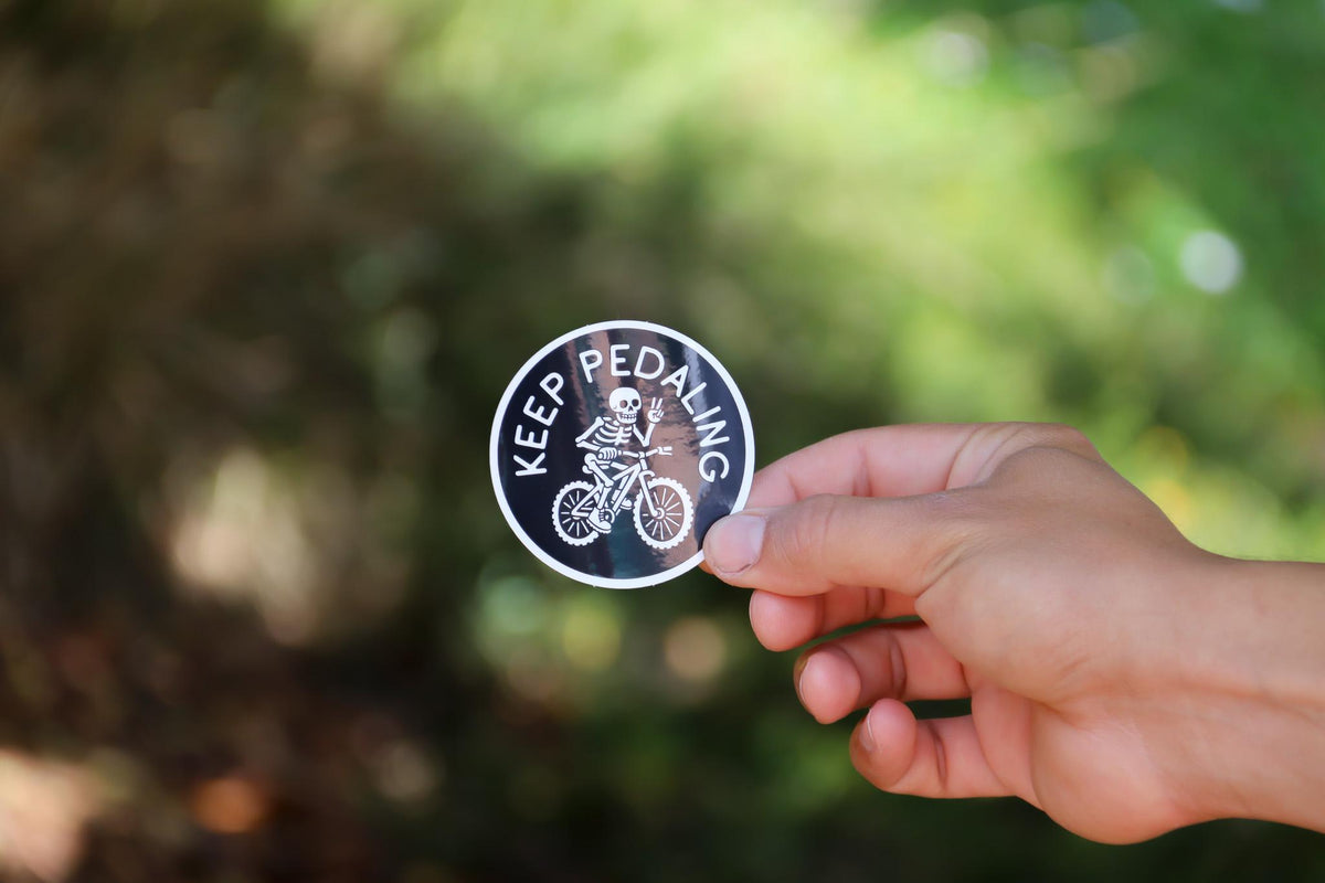 Sunlight catches the Keep Pedaling MTB sticker as a hand holds it along a shaded trail.