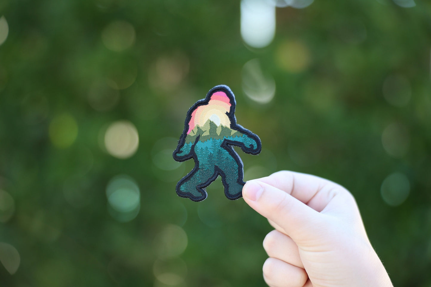 Held at arm's length against blurred green trees, the Bigfoot Adventure Patch looks small between finger and thumb.