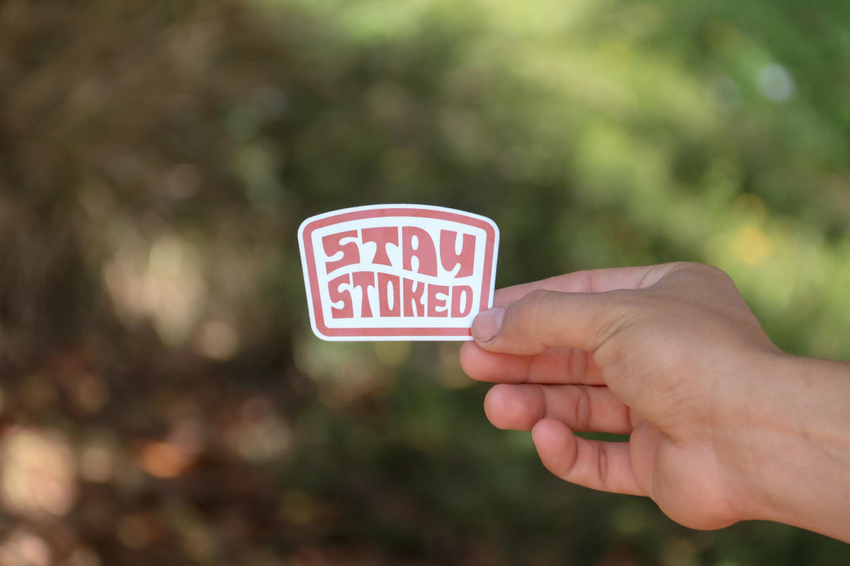 Out on a wooded trail, a hand extends the Stay Stoked sticker into dappled afternoon sun.