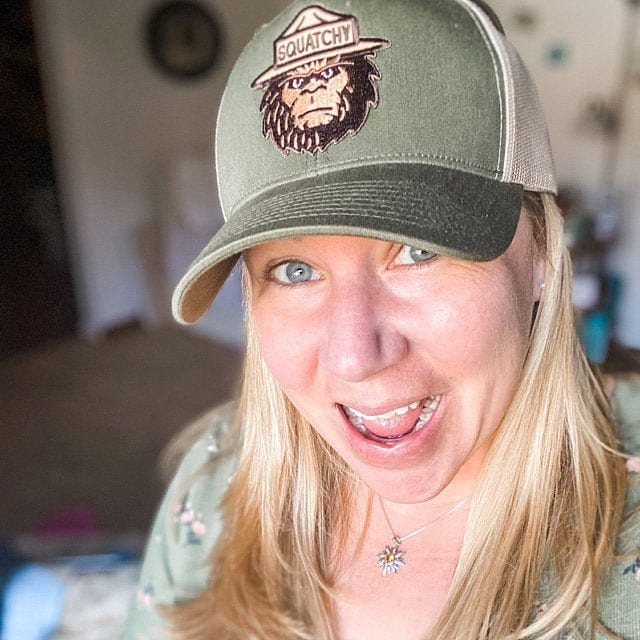 Smiling close up, a blonde woman wears the olive Bigfoot Squatchy trucker hat in a sunlit room.