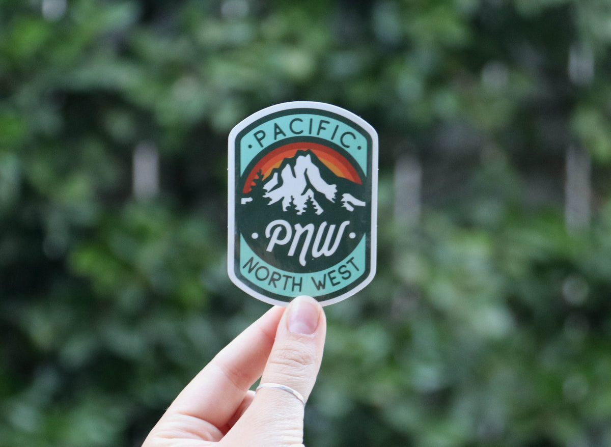 Wider view of a hand holding the PNW sticker, hedge filling the frame behind the mountain badge.