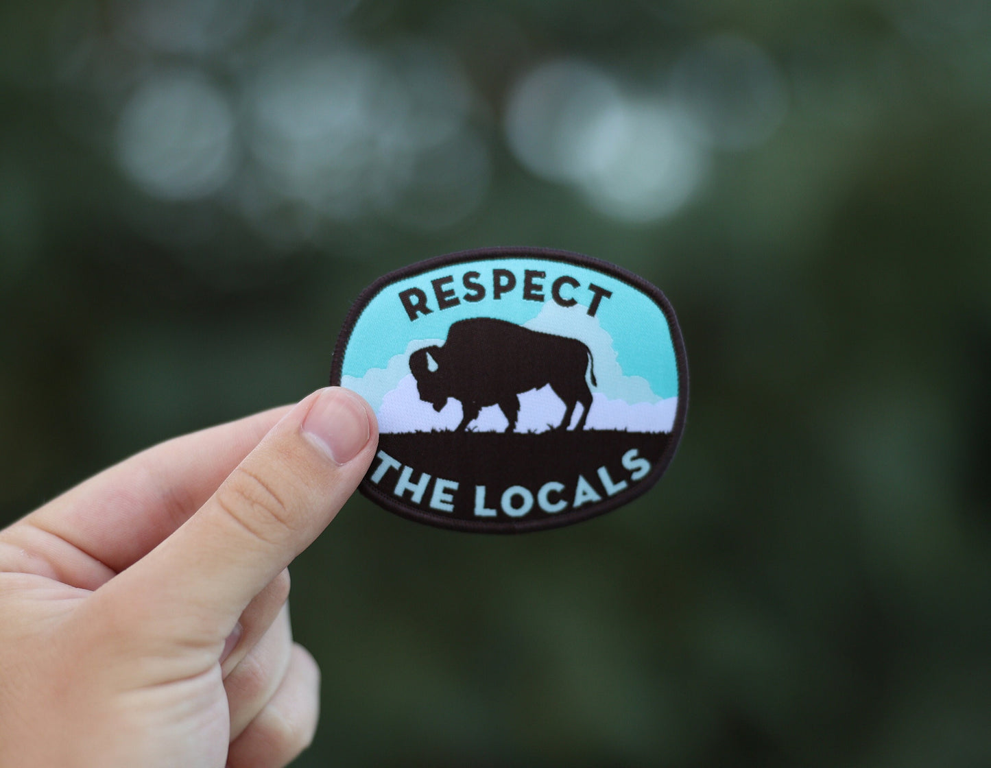 Fingers hold the Respect The Locals bison patch up against soft green trees thrown out of focus.