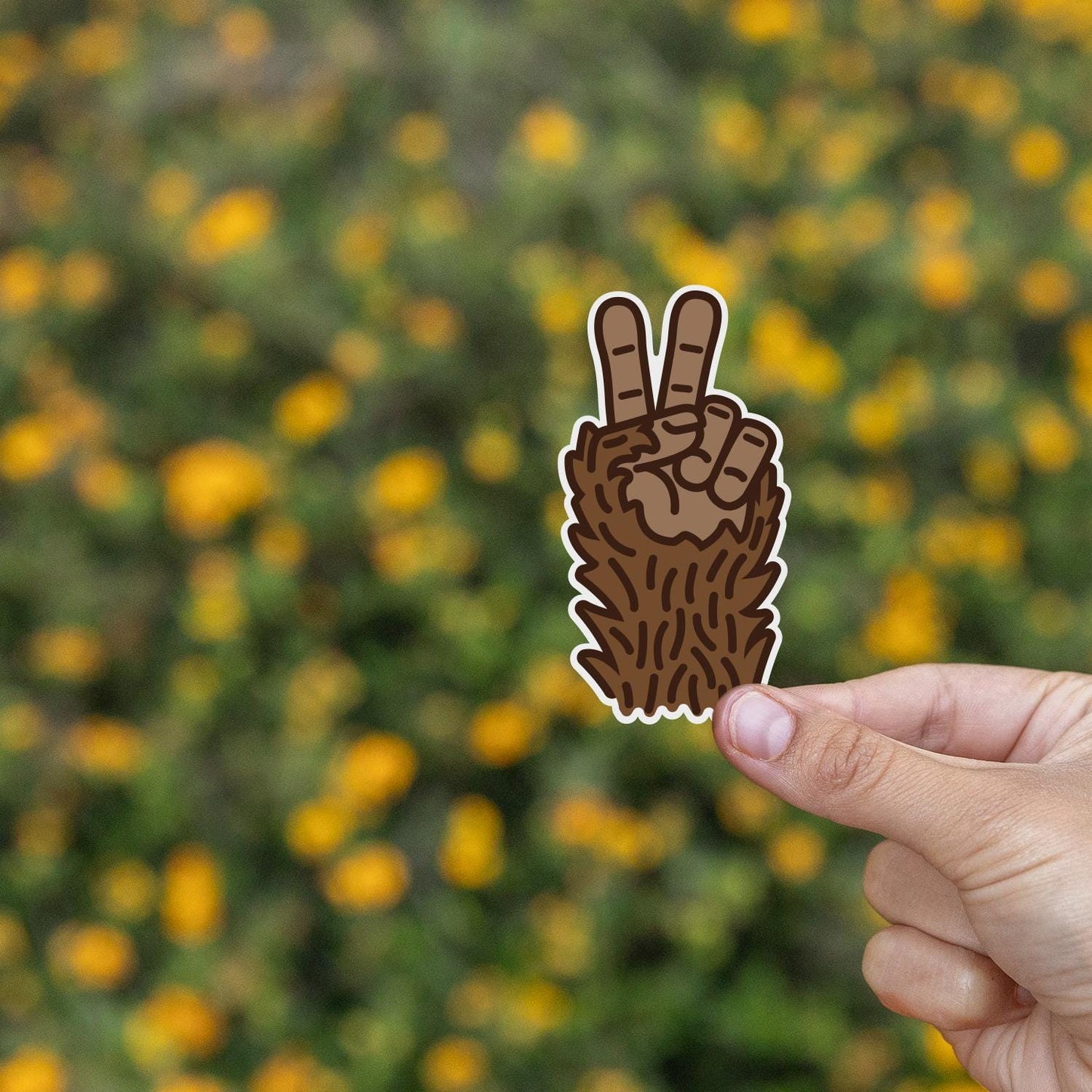 Yellow wildflowers fill the background while fingers hold the Bigfoot Peace sticker steady.