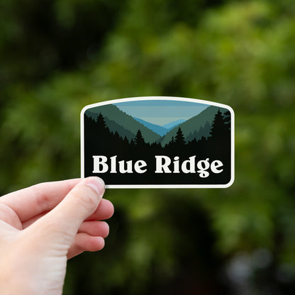Fingers pinch the Blue Ridge sticker in front of sunlit green foliage.