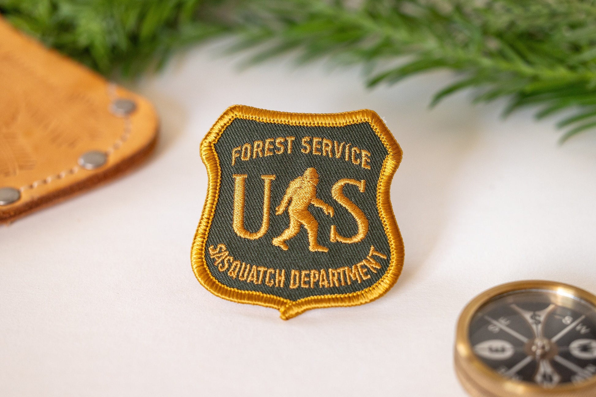 Beside a leather sheath and a brass compass, the US Sasquatch Department patch rests near evergreen sprigs.