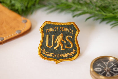 Beside a leather sheath and a brass compass, the US Sasquatch Department patch rests near evergreen sprigs.