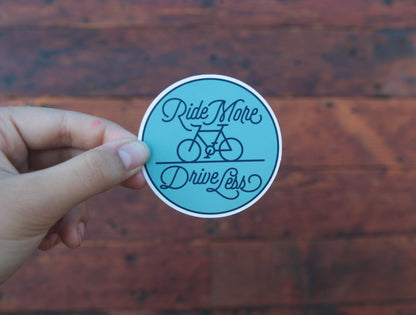 Pinched between finger and thumb, the Ride More Drive Less sticker stands out against dark wooden boards.