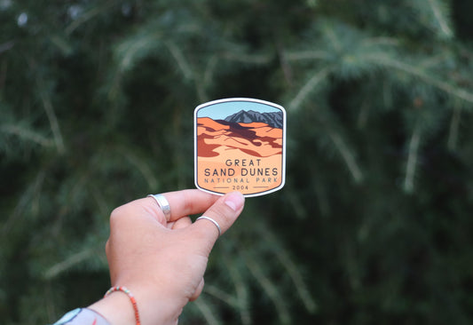 Held further back, the Great Sand Dunes National Park sticker shows its orange dunes and grey peak.