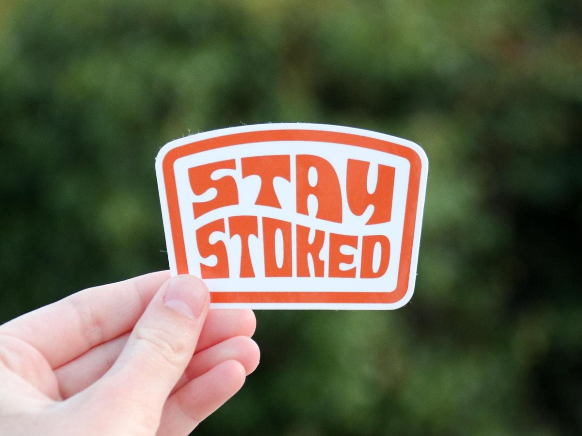 Turned slightly sideways, the Stay Stoked sticker catches soft light above dark green shrubs.