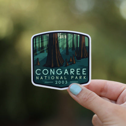 A hand with pale blue nail polish holds the Congaree National Park sticker against blurred green woods.