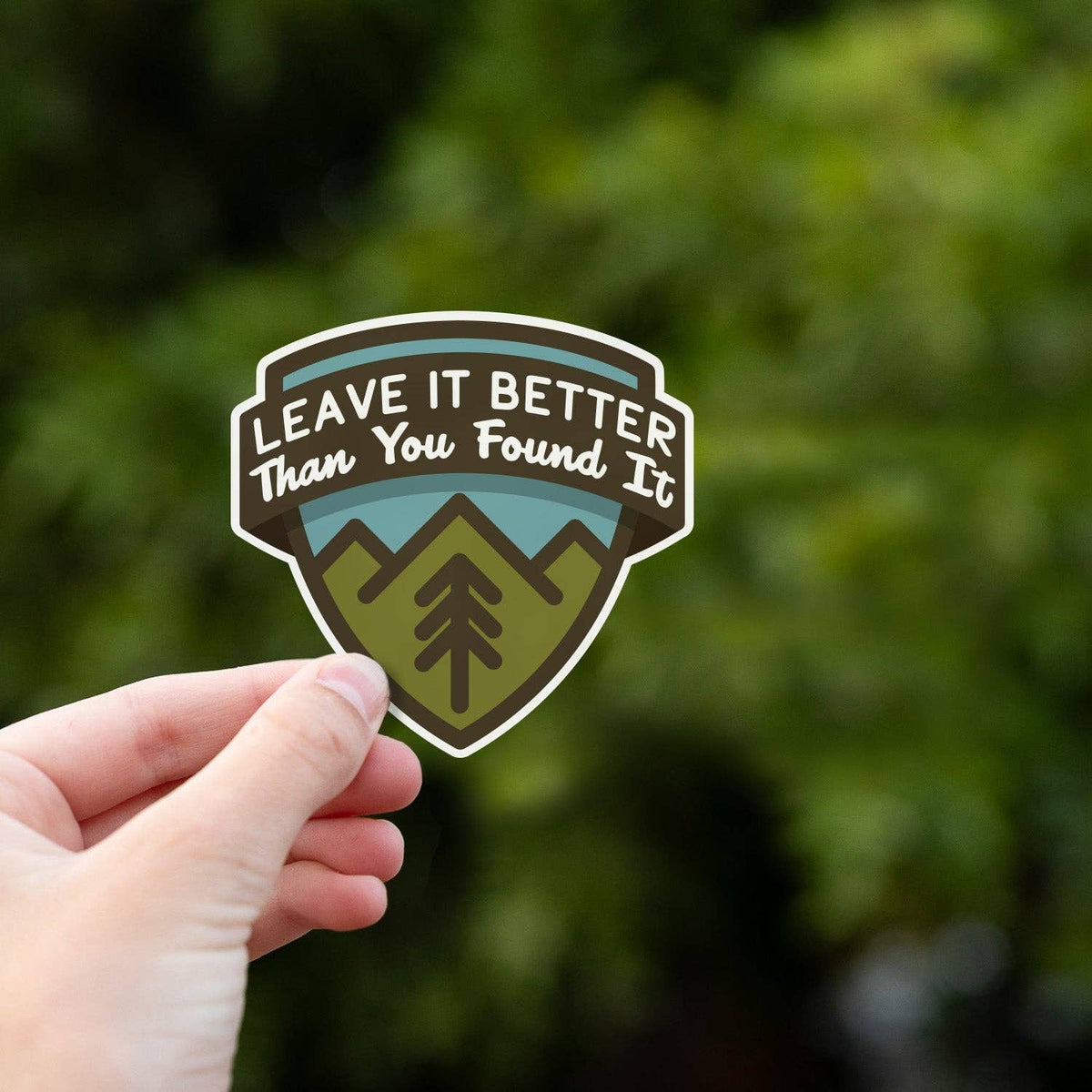 Held close to the camera, the Leave It Better Than You Found It sticker pops against blurred foliage.