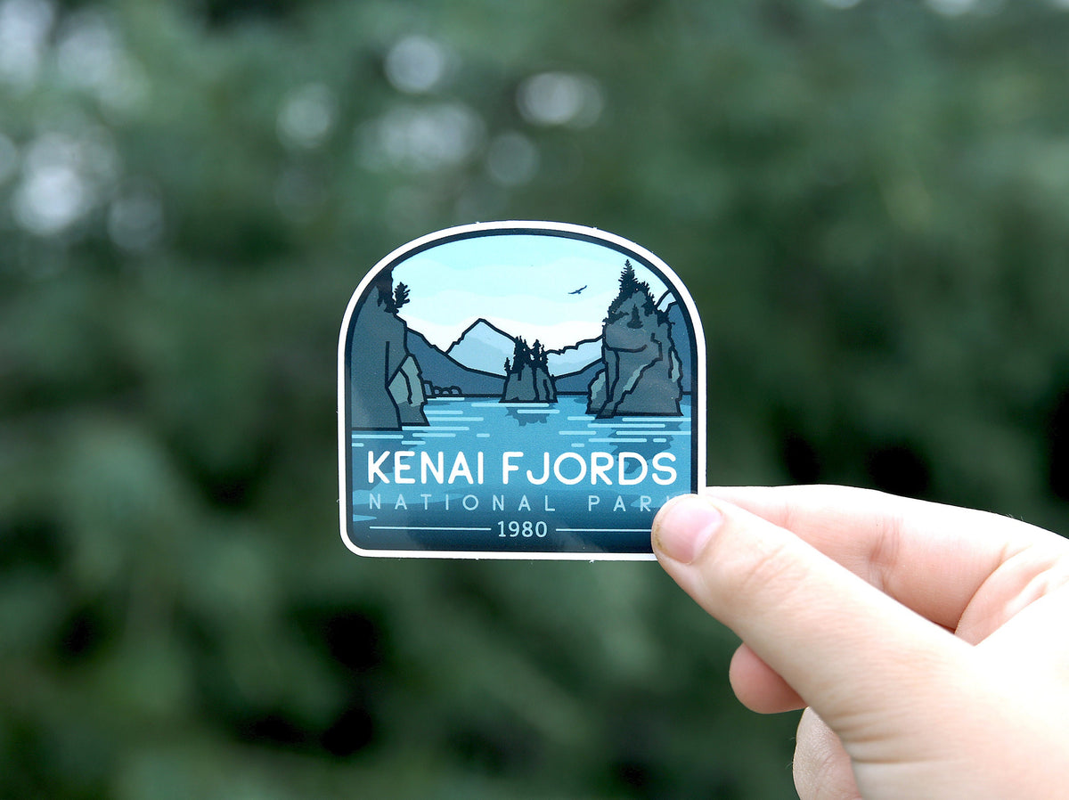 Held low in the frame, the Kenai Fjords National Park sticker catches daylight against a dense conifer.