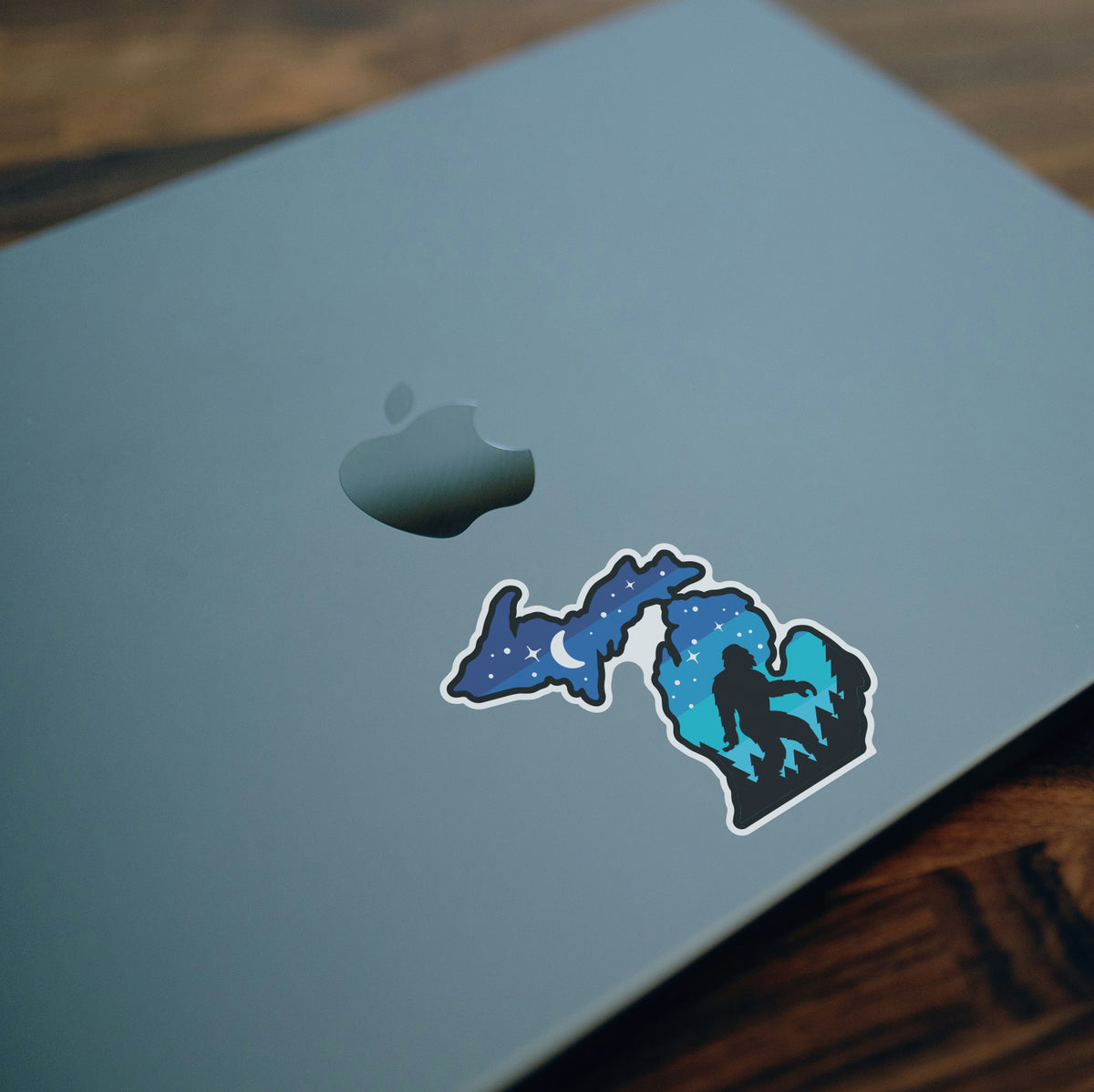 Placed below the Apple logo, the Michigan Sasquatch sticker decorates the lid of a closed laptop on a wood table.