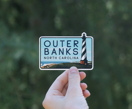 Framed against blurred green foliage, the Outer Banks North Carolina sticker sits smaller in the raised hand.