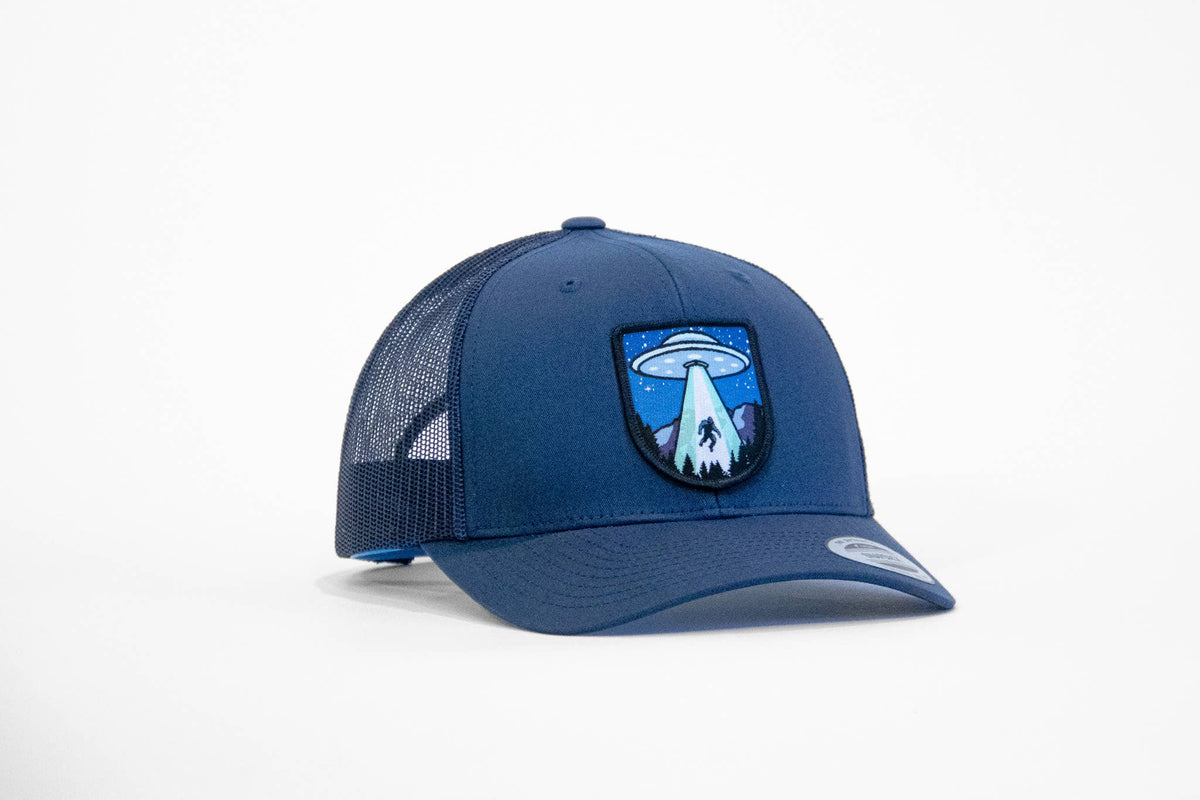 All navy Bigfoot UFO Conspiracy trucker hat turned slightly left, the flying saucer patch glowing over dark pines.