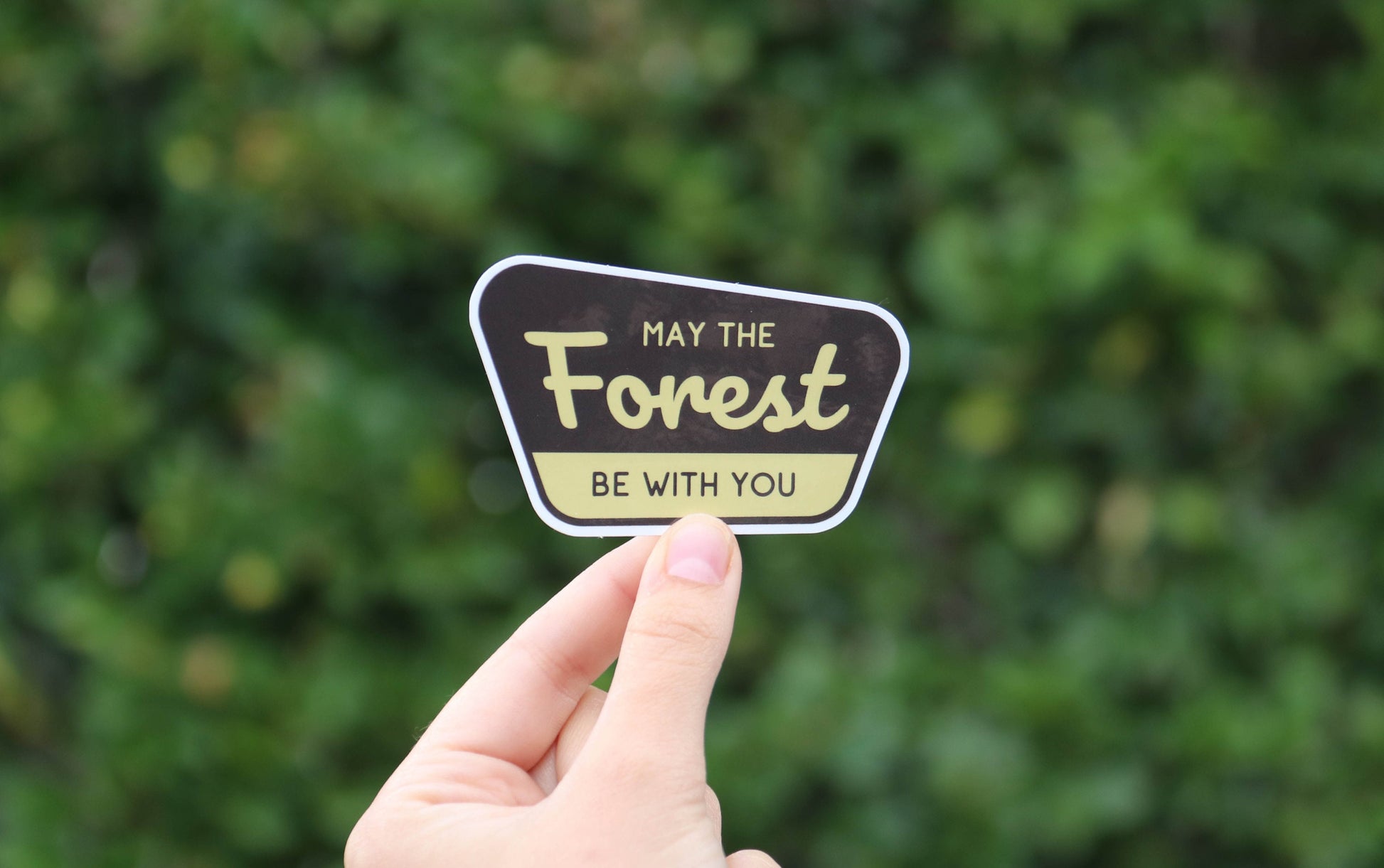 Two fingers lift the May The Forest Be With You sticker in front of a thick green hedge.