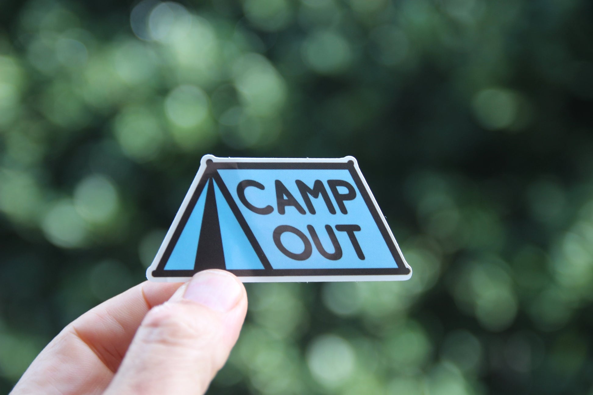 Held at arm's length, the Camp Out sticker catches sun on its glossy face with dark foliage behind.