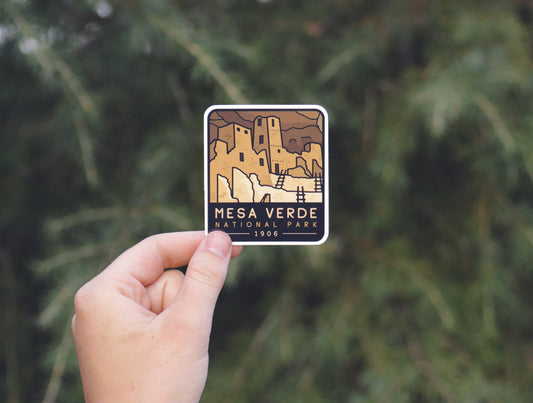 Held farther back against blurred evergreen branches, the Mesa Verde National Park sticker shows its 1906 date.