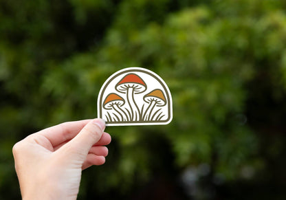 A hand holds the Three Mushrooms sticker up against blurred green leaves in soft daylight.