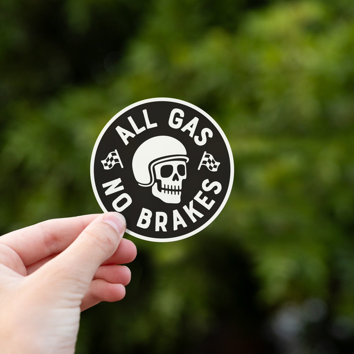 A hand holds up the All Gas No Brakes sticker against a backdrop of dense green leaves.
