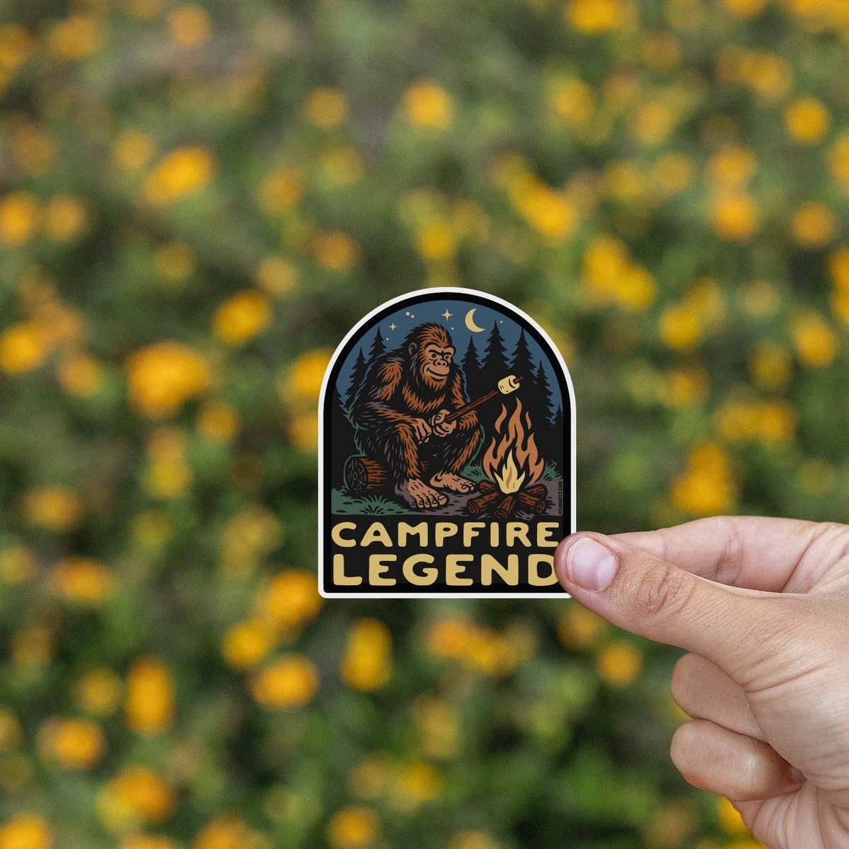 Held before a bank of yellow wildflowers, the Campfire Legend sticker glows with its fire and night sky art.