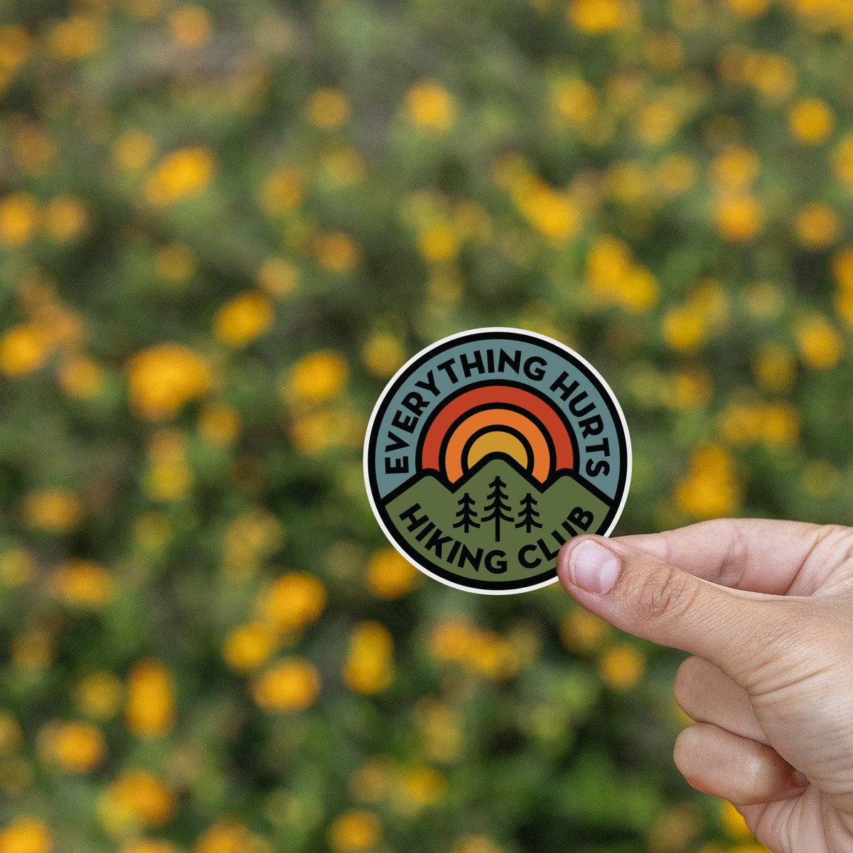 Yellow wildflowers blur behind the Everything Hurts Hiking Club sticker held between finger and thumb.