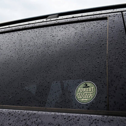 On the rain beaded window of a dark car, the Forest Witch sticker holds its place among the drops.
