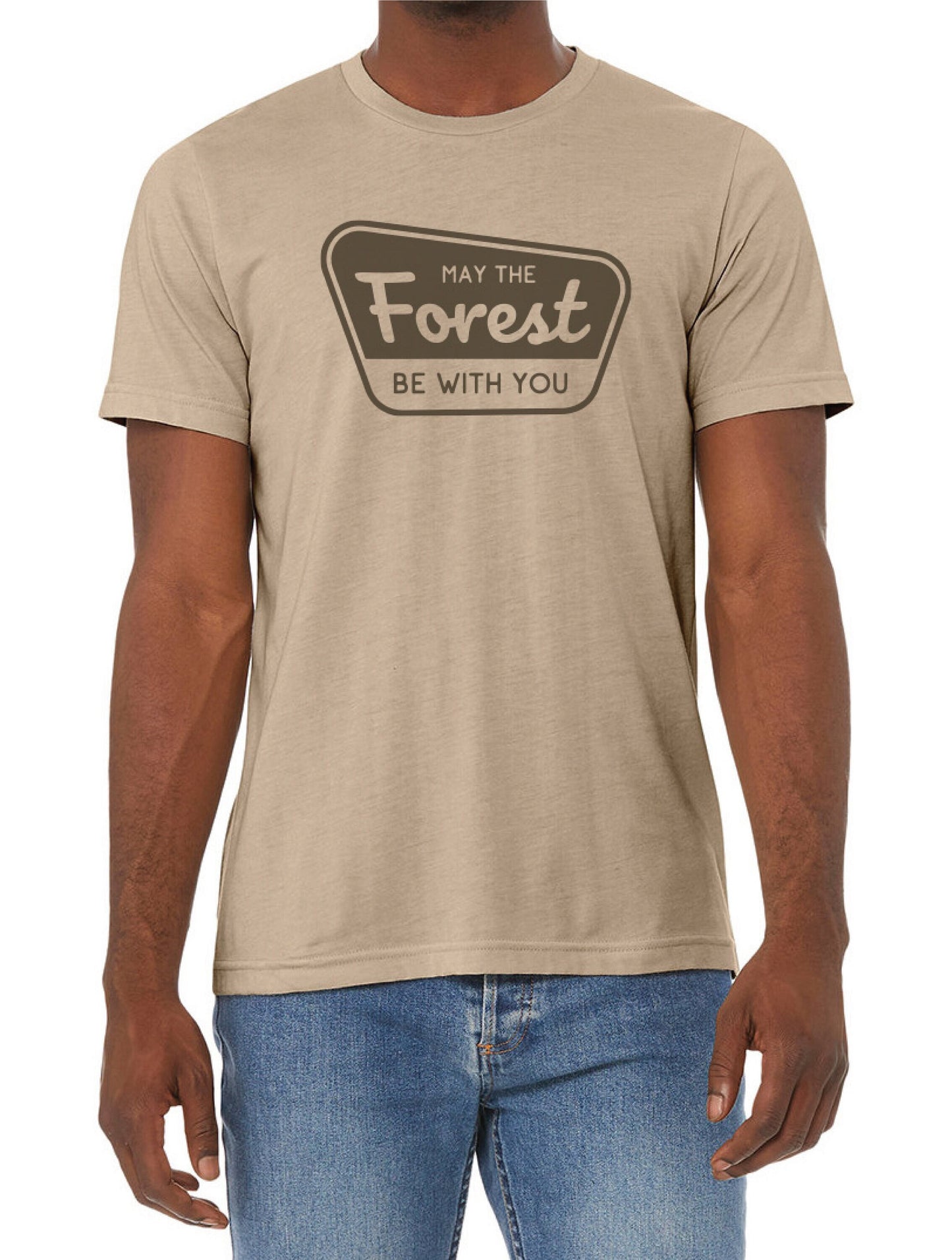 A man in blue jeans wears the tan May The Forest Be With You t shirt with its brown badge print.