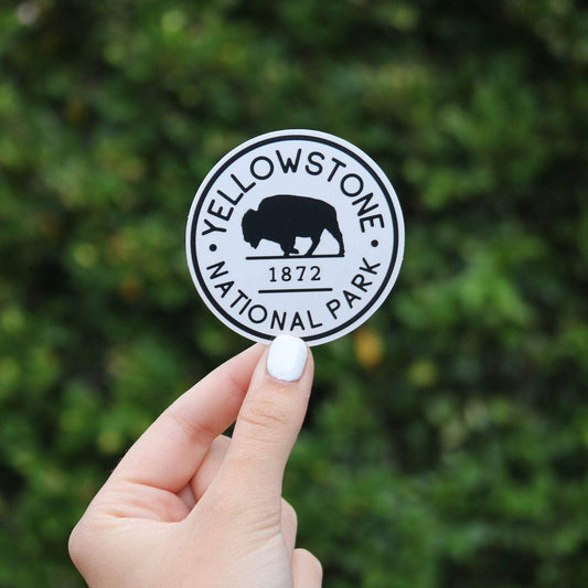 A hand holds the round black and white Yellowstone National Park sticker with its bison against a green hedge.