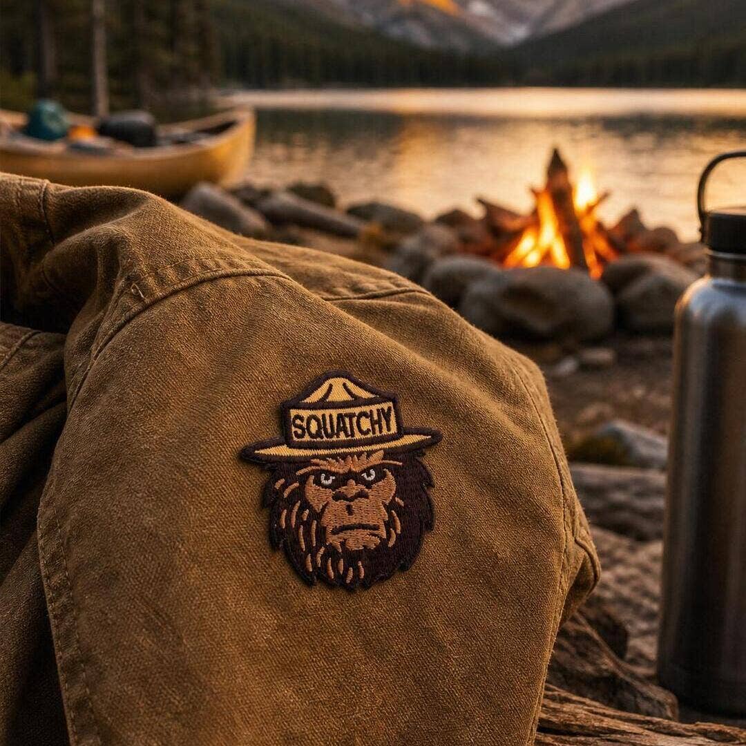Stitched onto the shoulder of a tan canvas jacket, the Squatchy patch sits near a lakeside campfire at dusk.
