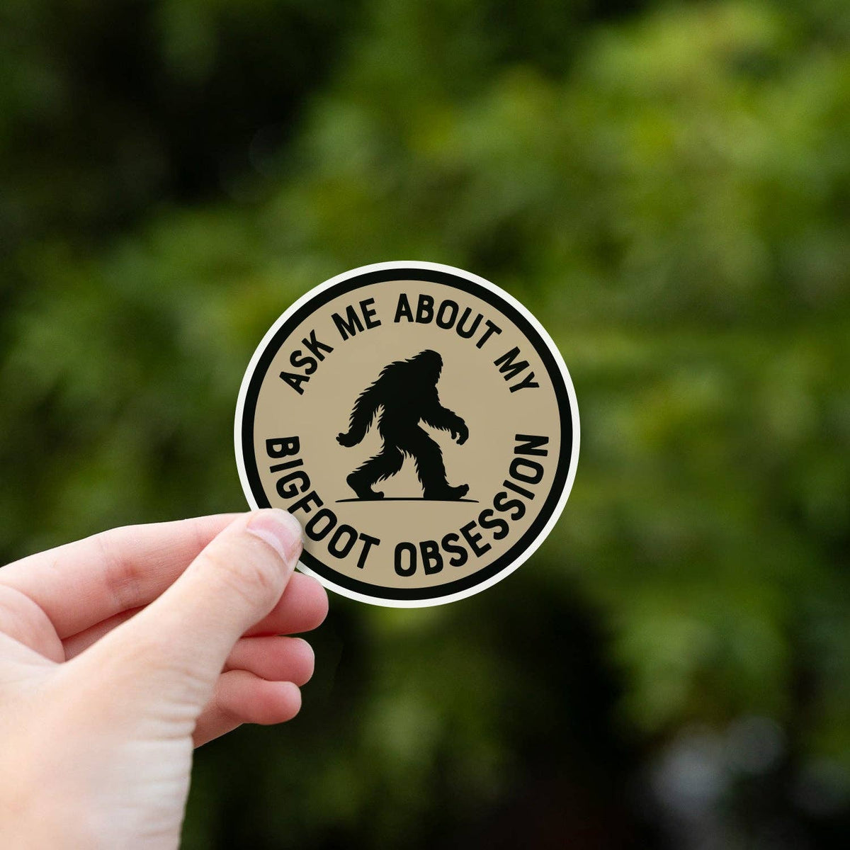 Close up, the Ask Me About My Bigfoot Obsession sticker fills the frame against soft green leaves.