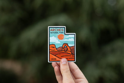 Fingertips lift the Arches National Park sticker in soft light, dark evergreen boughs falling out of focus.