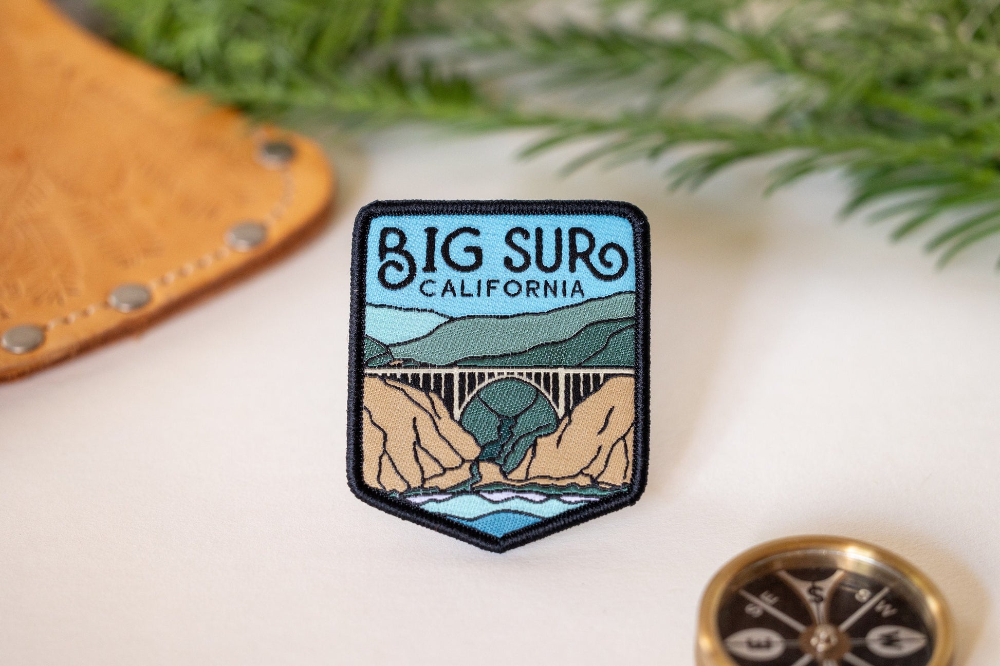 Beside a brass compass and a leather flap, the Big Sur California patch rests near sprigs of greenery.