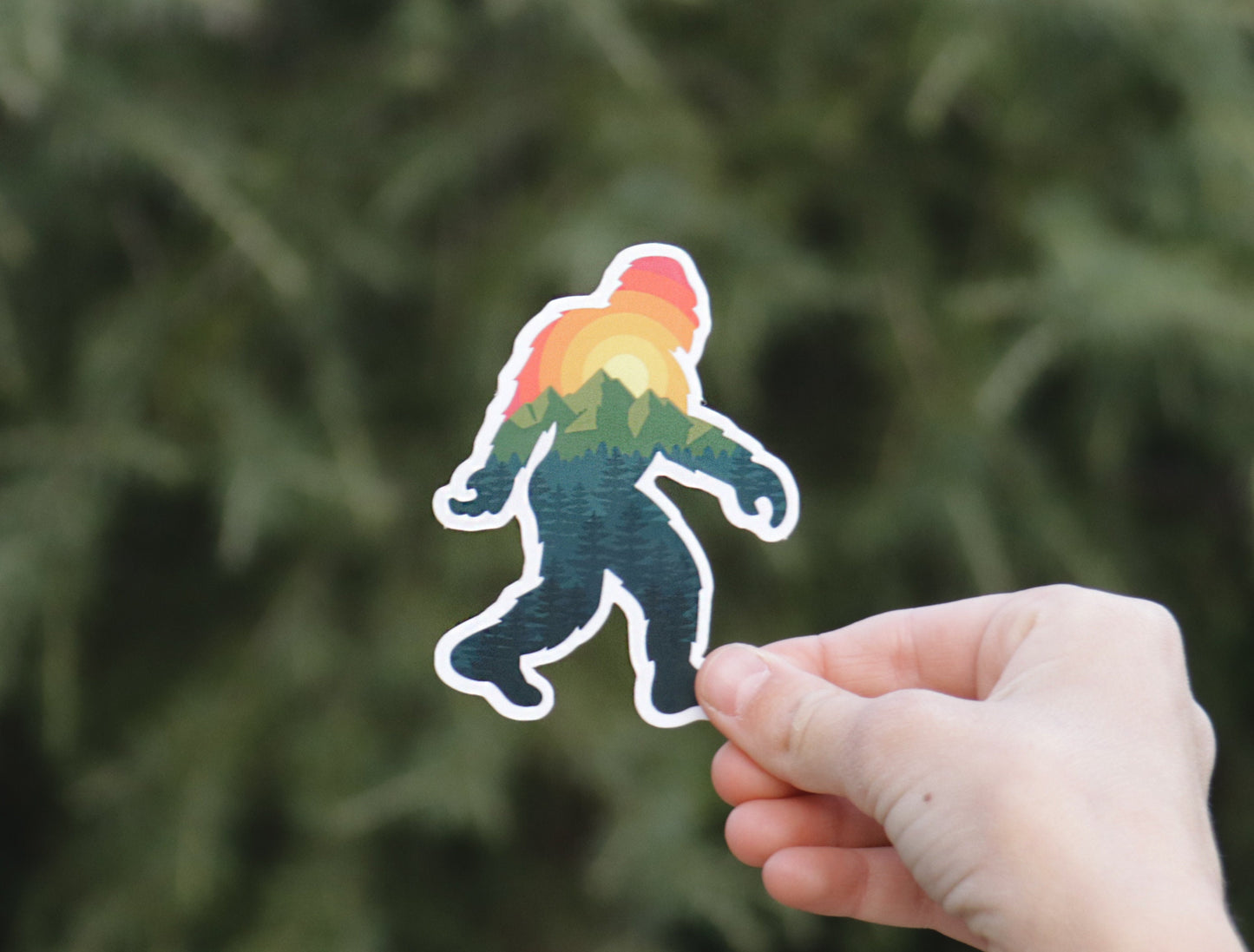 Framed wide against a hedge, the Bigfoot Adventure sticker rests between a thumb and forefinger.
