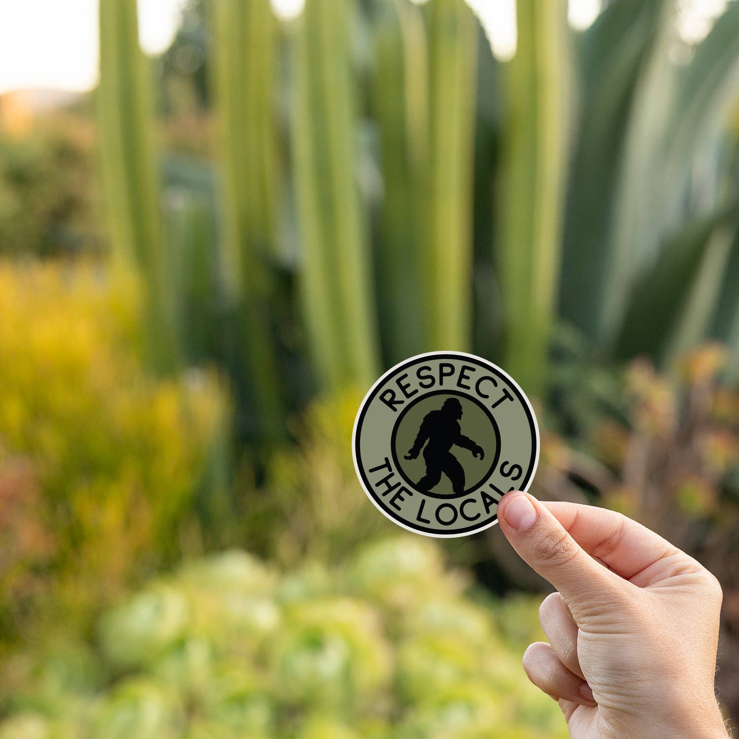 Tall cactus and succulents sit behind a hand holding the Bigfoot Respect The Locals sticker at golden hour.