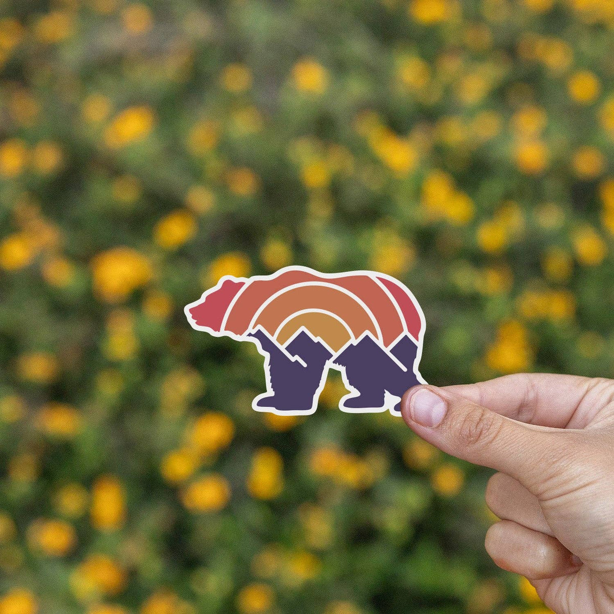 Yellow wildflowers blur behind the Adventure Grizzly Bear sticker pinched between two fingers.