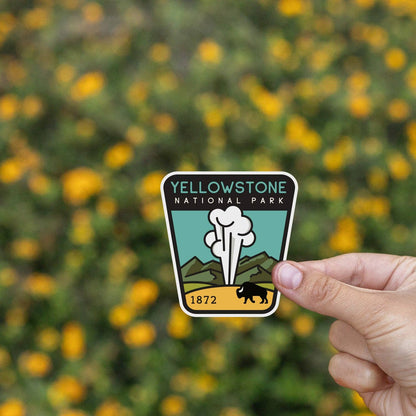 Fingers pinch the Yellowstone National Park sticker in front of a field of blurred yellow wildflowers.